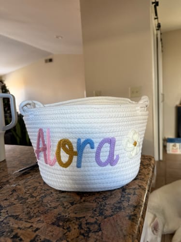 Customer's white personalized knitted basket with the name 'Alora' in multicolor yarn and a flower icon, sitting on a kitchen counter.