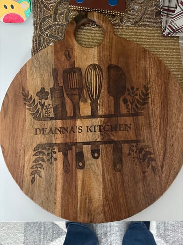 Customer's round wooden cutting board with a crisp, wood-burned design saying 'DEANNA'S KITCHEN' and showing kitchen utensils.