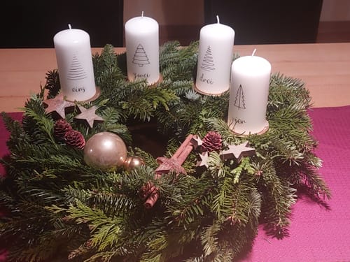 Ein Kunde zeigt die vier weißen Adventskerzen mit Baummotiven, die auf einem verzierten Adventskranz auf einem Tisch stehen.