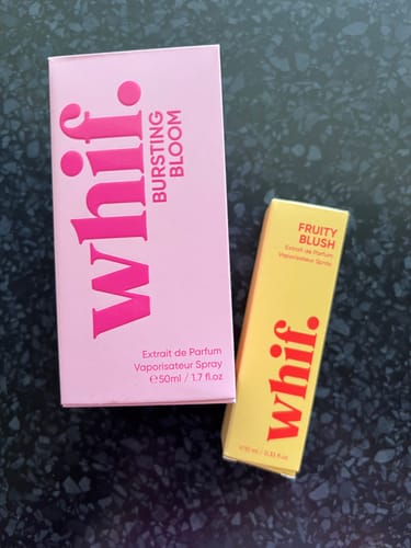 Reviewer's pink 'Bursting Bloom' and yellow 'Fruity Blush' perfume boxes on a dark surface.