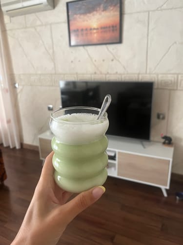 Mano de una clienta sosteniendo un Té Matcha Premium espumoso en un vaso de vidrio ondulado.