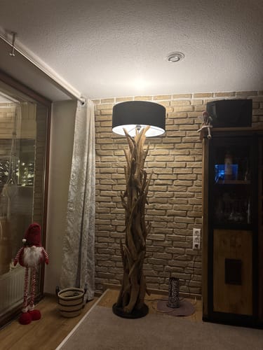 Ein Kunde zeigt die beleuchtete Stehlampe BLUMA aus Teakholz mit schwarzem Schirm in seinem gemütlichen Wohnzimmer.