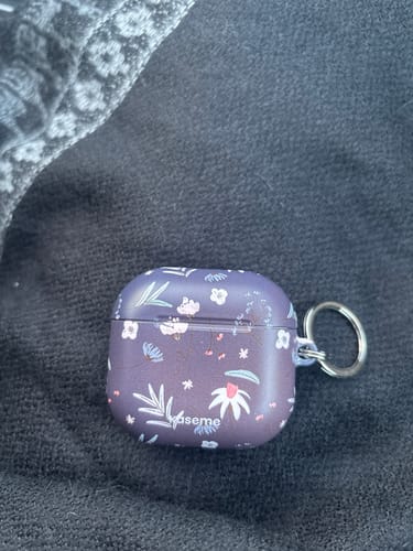 Reviewer's Mia Blue AirPods Case in a purple floral pattern, with a keychain ring attached, resting on a dark fabric surface.