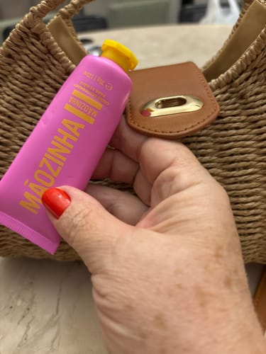 Cliente segurando o tubo rosa do creme Mãozinha na frente de uma bolsa de palha.