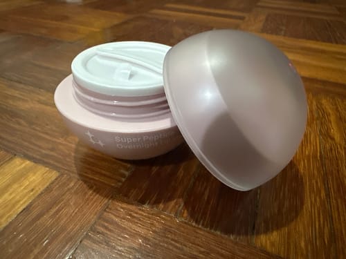 Customer's open pink jar of Super Peptide Overnight Lip Mask with the lid beside it on a wooden surface.