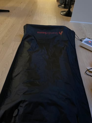 Anmelders sorte infrarøde saunatæppe er lagt ud på et trægulv med sin kontrolenhed ved siden af.