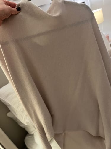 Customer holding up the light beige Asymmetric Draped sweater, showing the soft seamless knit material indoors.