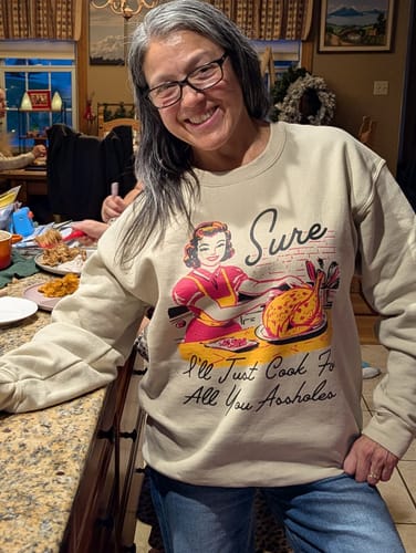 Reviewer smiling while wearing the beige 'Sure I'll Just Cook For All You' sweatshirt at a family dinner.