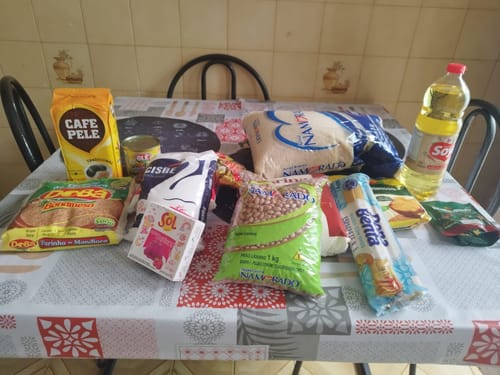 Cliente mostrando os itens da Cesta Básica Pinus Plus, como arroz, feijão e café, espalhados sobre uma mesa.