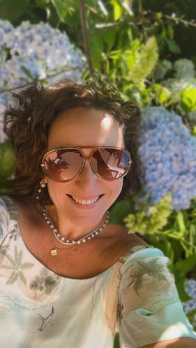 Cliente sorrindo e usando os Óculos Veneza oversized, deitada em um jardim com flores azuis ao fundo.