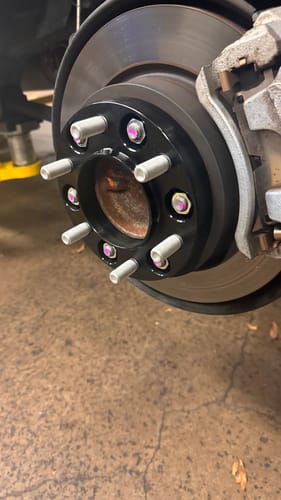 Close-up of a customer's black 1-inch forged wheel spacer installed on the wheel hub of a vehicle in a garage.