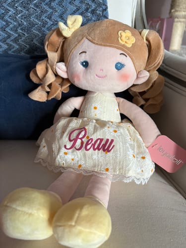 A customer's adorable Personalized Daisy Girl doll sitting on a chair, showing the name 'Beau' embroidered on its yellow dress.