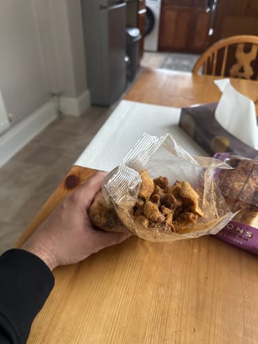 Customer holding an open bag of crunchy Salt & Cracked Black Pepper Pork Scratchings over a wooden table.
