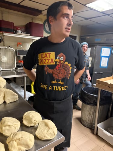 Customer wearing the black Eat Pizza Save A Turkey T-Shirt in a kitchen next to pizza dough.