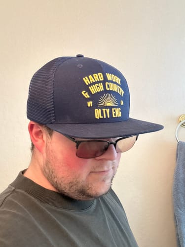 A reviewer wearing the navy blue Big Tall Trucker Hat with a flat brim and yellow lettering.