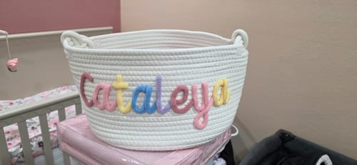Reviewer's white knitted basket with the name 'Cataleya' in multicolor yarn, shown in a nursery setting.