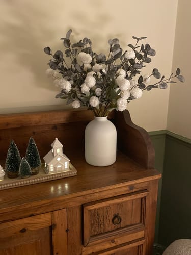 Customer's Faux Arrangement of white flowers and green eucalyptus shown in a white vase on a wooden cabinet.
