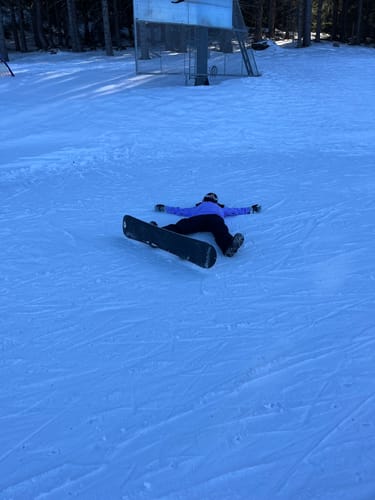 Reviewer in baggy snow pants lying on a snowy slope next to their snowboard, showing the fit.