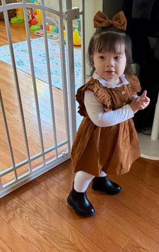Customer's toddler wearing the rust Serenity Corduroy Dress with the matching bow while standing on a wood floor.
