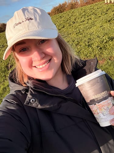 Reviewer smiling outdoors, wearing the Pilates & Lattes Cap and holding a cup of coffee.