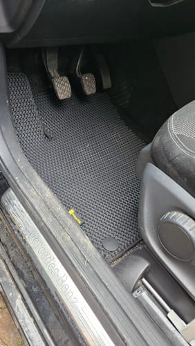 Recenzent pokazuje czarny, dopasowany dywanik samochodowy po stronie kierowcy, przy pedałach.