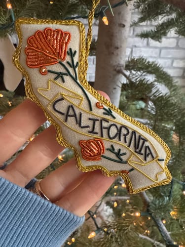 Reviewer holding the California-shaped Poppy Beaded Ornament, showing the detailed embroidery and gold trim.