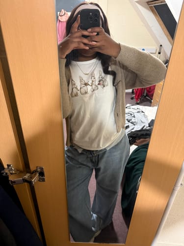Reviewer taking a mirror selfie wearing the all that jazz baby tee with a cardigan and jeans.