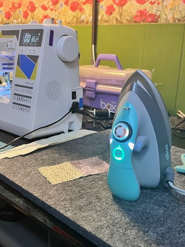 Reviewer's teal Oliso Mini M3Pro Project Iron on a sewing table next to a sewing machine.