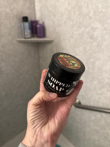 Customer holding a black jar of Whipped Soap in their hand inside a shower.