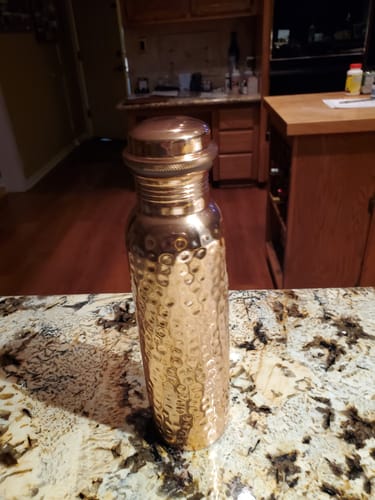 Reviewer's shiny, hammered Himalayan Ayurvedic Pure Copper Bottle sitting on a granite kitchen countertop.