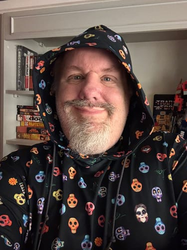 Reviewer smiling while wearing the black Coco "Calavera" hoodie with its colorful pattern and the hood up.