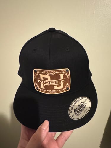 Customer holding a black custom monogram hat. The leather patch is engraved with large 'NJ' initials over the name 'NOBLE'.