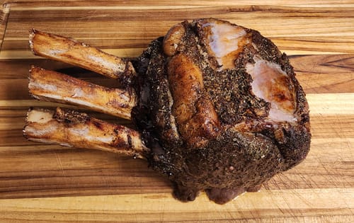 Reviewer's cooked Tomahawk Prime Rib Roast with a dark herb crust, resting on a wooden cutting board.