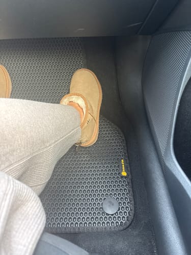 Recenzent pokazuje czarny, dopasowany dywanik samochodowy z fakturą plastra miodu w swoim aucie.