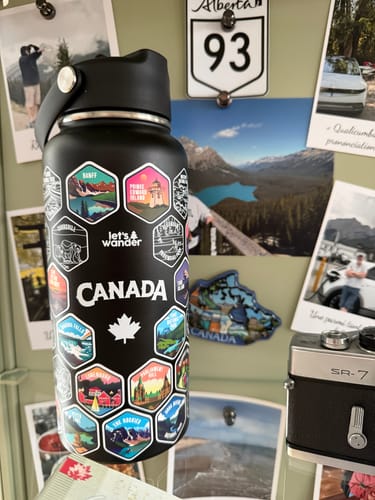 Reviewer's black Canada Bottle, covered in colorful destination stickers, in front of a bulletin board with photos.