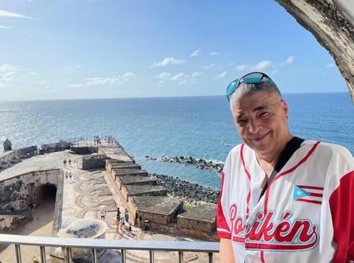 Customer in a white and red Puerto Rico Heritage Custom Baseball Jersey, smiling with a coastal fort in the background.
