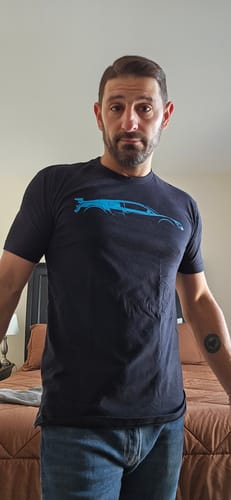 Reviewer wearing the dark 80Eighty® Mystery Shirt with a blue car graphic, showing the fit in a bedroom.