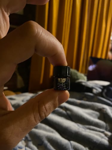 A reviewer holds a tiny vial of Twilight Maze fragrance between their fingers.