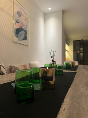 Recensent's brandende Vanilla Noir Geurkaars in een gouden pot op een eettafel met groen glaswerk.