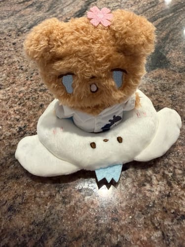 Customer's brown Younghoon Tanningdoll with teary eyes and a pink flower on its head, sitting on a white plush.