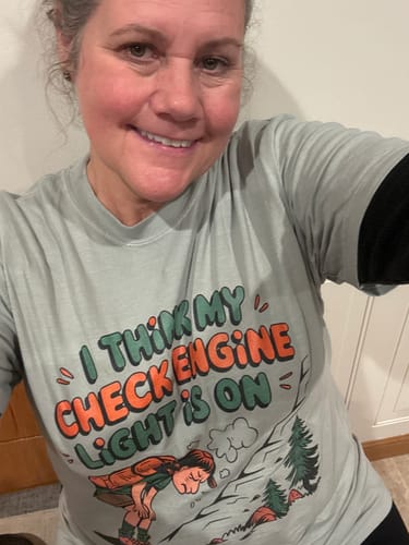 Reviewer smiling in a light green Check Engine Light T-shirt with a funny tired hiker graphic.