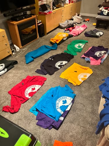 Customer's colorful t-shirts on a floor, each with a white logo made from V2 Pro Heat Transfer Film.