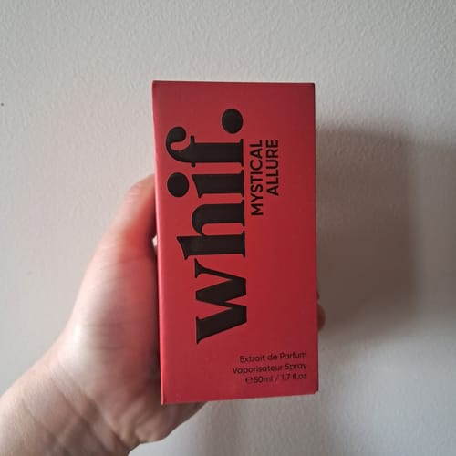 Customer's hand holding the red box of Mystical Allure perfume against a white wall.
