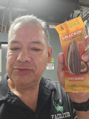 Reviewer in a work shirt holding up a bag of Smackin' Limited Release Spicy Queso sunflower seeds.