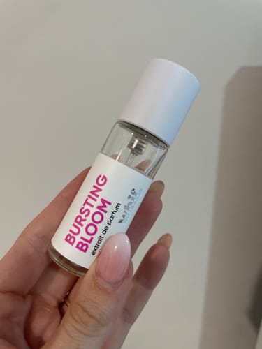 Customer's hand holding a small, clear bottle of Bursting Bloom perfume against a plain background.
