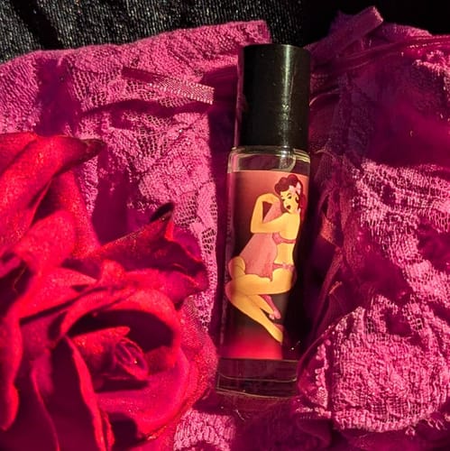 Customer's Afterglow perfume bottle, with its pin-up label, displayed on pink lace next to a red rose.