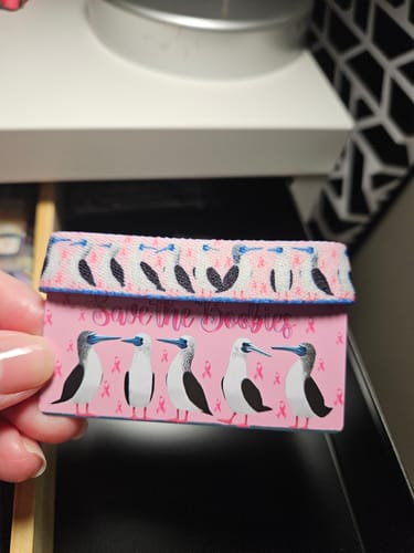 A reviewer holds the Save the Boobies Bracelet wrapped around its pink card, which features blue-footed booby birds.