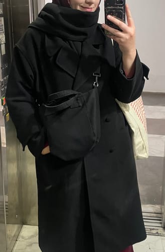 Yorumcu, oversize atkılı siyah kaşmir kabanı giyerken ayna karşısında poz veriyor.