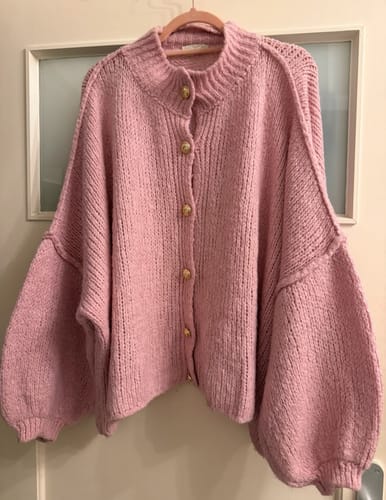 Kunde zeigt den übergroßen, rosa 'CozyGlam' Cardigan mit goldenen Knöpfen, der an einem Kleiderbügel hängt.
