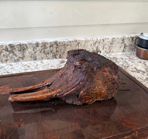 Customer's cooked Tomahawk Prime Rib Roast with a dark crust resting on a wooden cutting board in a kitchen.